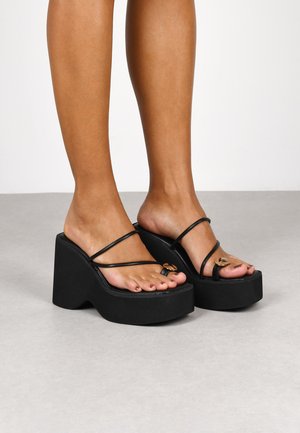 Sandalias de plataforma negras con tiras delgadas, que presentan un acento metálico decorativo y una suela de plataforma texturizada.