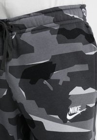 Grå kamouflage joggbukser med dragsko i midjan, tillverkade av mjukt material, med en vit Nike-logotyp på vänster sida.