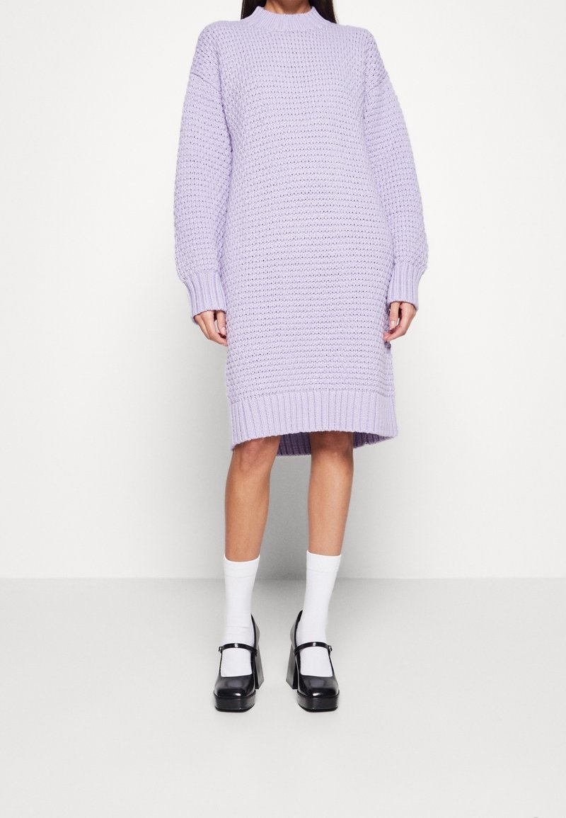 Robe en tricot lilas avec un col montant, des manches surdimensionnées et des ourlets côtelés. Portée avec des chaussettes blanches et des chaussures compensées noires.