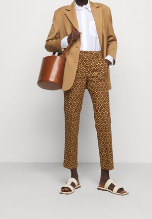 Personne portant un blazer camel, une chemise blanche, un pantalon court à motifs, des sandales blanches, et tenant un sac seau en cuir marron à l'épaule.