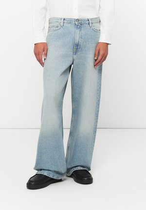 Person wearing light blue wide-leg jeans, black shoes, and a white long-sleeve shirt standing against a plain white background.