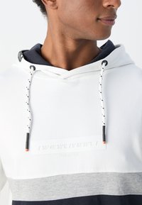 Felpa con cappuccio di colore bianco nella parte superiore, grigia nel mezzo e blu navy nella parte inferiore. Presenta dei lacci a contrasto e un'etichetta testurizzata con le coordinate.