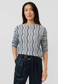 Pullover mit welligem marineblauem und cremefarbenem Muster, 3/4-langen Ärmeln und einem Bootsausschnitt, kombiniert mit hochtaillierter dunkler Hose und silbernem Gürtel.