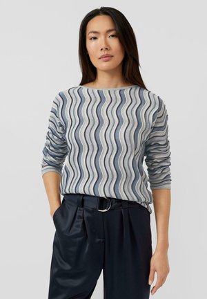 Pullover mit welligem marineblauem und cremefarbenem Muster, 3/4-langen Ärmeln und einem Bootsausschnitt, kombiniert mit hochtaillierter dunkler Hose und silbernem Gürtel.