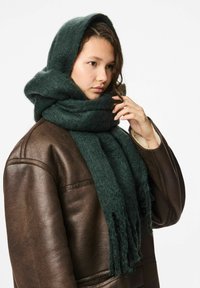 Scarf verde e soffice realizzato in materiale morbido, drappeggiato come cappuccio, con bordi frangiati. Indossato sopra una giacca di pelle marrone.