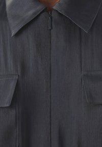 Detailní záběr na tmavě šedou texturovanou bundu s límcem, předním zipem a dvěma klopovými kapsami po obou stranách.