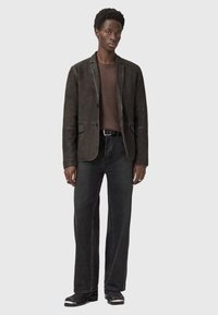 Uomo che indossa un blazer in suede marrone scuro, un maglione marrone, una cintura nera, jeans grigio scuro e stivali neri, in piedi contro uno sfondo semplice.