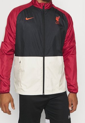Mand iført en rød, sort og hvid Liverpool FC Nike jakke med lynlås, kombineret med sorte Nike-shorts.