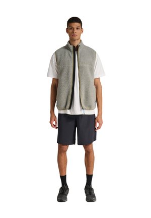 Jeune homme debout portant un gilet polaire gris, une chemise blanche à manches courtes, un short noir, des chaussettes noires et des baskets noires sur fond blanc.