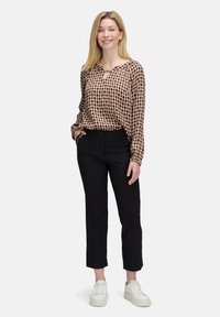 Vrouw staat, draagt een gestreepte blouse met lange mouwen, zwarte broek met ritszakken en witte platform sneakers, en glimlacht naar de camera.