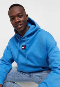 Felpa blu in cotone, con un piccolo logo ricamato in rosso, bianco e blu. Il design include una tasca frontale e cappuccio con cordini.