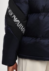 Mornarska puffer jakna s crnim panelom i bijelim "Emporio Armani" logotipom. Tekstura je glatka i blago sjajna s kviltanim dijelovima.