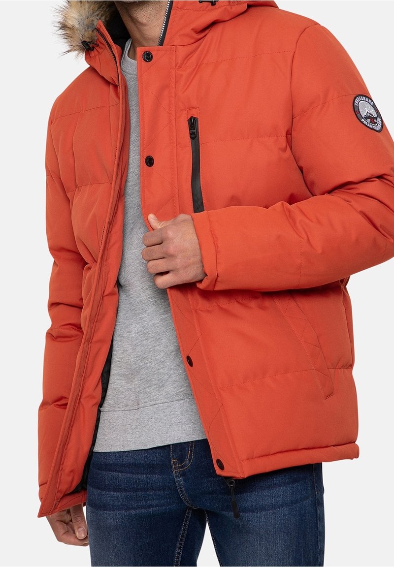 Homme portant une veste matelassée orange avec une capuche bordée de fourrure, un pull gris et un jean bleu, ajustant la fermeture éclair de la veste.