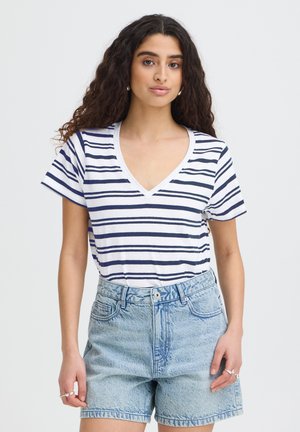 Mlada žena s dugom kovrčavom kosom odjevena u bijelu i mornarsko plavu prugastu majicu s V-izrezom i svijetlo plave traper kratke hlače.