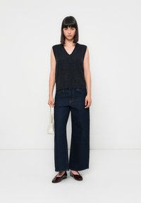 Donkergrijze mouwloze V-hals gebreide top gecombineerd met wijde donkere blauwe jeans. Het model houdt een kleine witte handtas vast en draagt bordeauxrode flats.