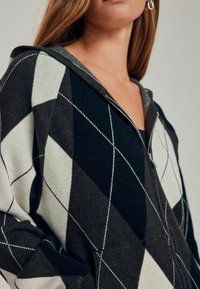 Bluza z kapturem na zamek o diamentowym wzorze w ciemnozielonym, kremowym i czarnym kolorze. Posiada fakturowany materiał oraz kontrastujące szwy.