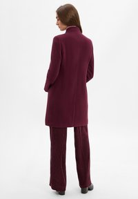 Burgunderfarbener Mantel mit hohem Kragen und gerader Silhouette, kombiniert mit ribbeligen burgunderfarbenen Hosen, die eine glatte Textur und minimale Details aufweisen.