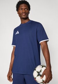 Μπλε-ναυτικό αθλητικό μπλουζάκι με κοντά μανίκια, λευκή επένδυση και λευκό λογότυπο στο αριστερό στήθος. Μαλακό ύφασμα, άνετη εφαρμογή, φορεμένο με μπάλα ποδοσφαίρου.