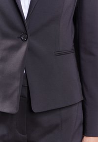 Blazer noir sur mesure avec une texture lisse, fermeture à un bouton, petit revers en pointe discret et poches latérales, assorti à un pantalon coordonné.