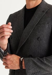 Grijze dubbelzijdige blazer van een wolmix met zwarte knopen, voorzien van een borstzak en een gestreepte voering. Decoratieve armbanden om de pols.