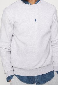 Sudadera gris claro con cuello y puños acanalados. Presenta un pequeño logo azul marino en el pecho y se superpone a una camisa de mezclilla por debajo.