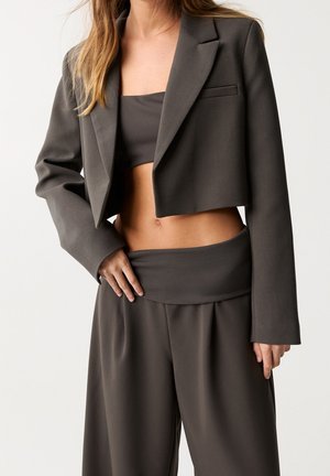 Mujer que lleva un blazer corto oscuro, top bandeau a juego y pantalones de pierna ancha de talle alto, posando con una mano cerca de la cintura.