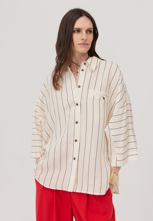 Kobieta w luźnej kremowej bluzce z czarnymi pionowymi paskami i czerwonych spodniach, stojąca z jedną ręką w kieszeni na jednolitym tle.