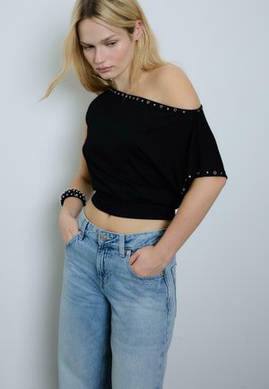 Femme blonde portant un haut noir à épaules dénudées avec des œillets et un jean bleu clair taille haute, les mains dans les poches, regardant vers le bas.