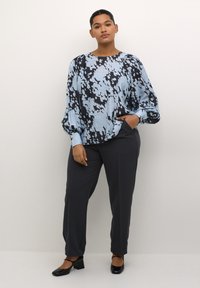 Luftige hellblaue Bluse mit schwarzem abstraktem Muster und puffierten Ärmeln; kombiniert mit dunkelgrauen, taillierten Hosen und schwarzen High Heels.
