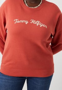 Personne portant un sweatshirt rouge Tommy Hilfiger et un jean bleu, accessoirisé d'un collier de perles en marguerite blanches et jaunes.