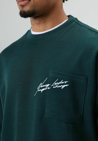Dunkelgrüner Pullover mit rundem Halsausschnitt, Brusttasche und weißer bestickter Schrift. Verfügt über eine weiche Stoffstruktur und ein lässiges Design.