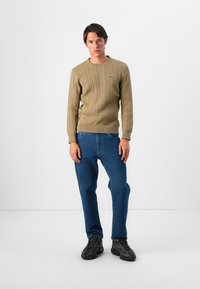 Pull en tricot cable beige avec des poignets et un ourlet côtelés, associé à un jean en denim bleu et des baskets noires. Design simple, surface texturée.