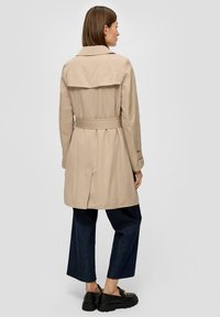 Beige trenchcoat met een ceintuur in de taille, een dubbelzijdig ontwerp en een opening aan de achterkant. Gestyled met donkerdenim wijde broek en schoenen.