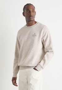 Világos bézs pulóver, puha anyagból készült, kerek nyakkivágással és bordázott mandzsettákkal rendelkezik. Sötétvörös nyomtatott "TOMMY HILFIGER" logó található rajta.
