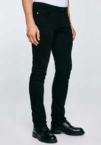 Mężczyzna w czarnych dopasowanych spodniach i czarnych skórzanych butach sznurowanych, stojący na tle jasnego tła.