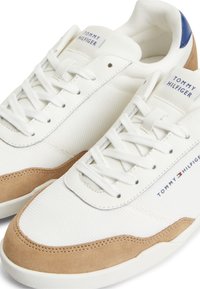 Άσπρα αθλητικά Tommy Hilfiger με ταμπά σουέτ λεπτομέρειες, ύφασμα από δίχτυ, μπλε ταμπελάκι στη φτέρνα και λογότυπα στην γλώσσα και πλευρά.
