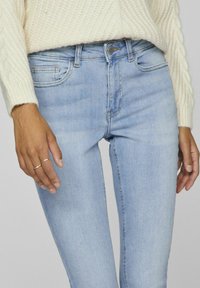 Vaqueros de mezclilla azul claro con un corte ajustado, diseño de cinco bolsillos y costuras sutiles. Combinados con un suéter de punto trenzado color crema.