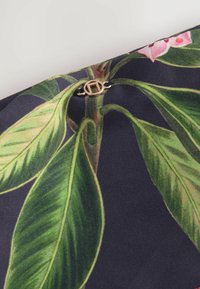 Blommönstrad tyg i mörka färger med gröna blad och rosa blommor, accentuerad av en cirkulär detalj i guld.