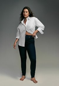 Camisa blanca de botones parcialmente metida en unos jeans skinny azul marino, mostrando un ajuste relajado y las mangas arremangadas. El modelo está descalzo.