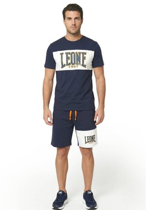 Un homme porte un T-shirt Leone bleu marine et un short assorti, avec des empiècements blancs ornés du logo et des cordons orange, sur un fond blanc uni.