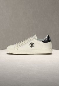 Baskets basses en cuir blanc avec languette de talon noire, lacets blancs et logo métallique décoratif sur le côté.