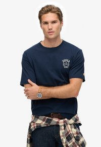 Joven con cabello corto rubio, vestido con una camiseta de Superdry azul marino, una camisa de cuadros atada alrededor de la cintura, jeans oscuros y un reloj de pulsera.