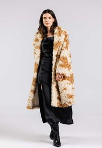 Faux fur-kappa i krämfärgade och karamellmönster, knälång, oversized passform, kombinerad med en svart satin-klänning med en sidoklyvning och svarta stövlar.