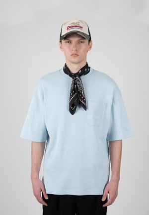 Mladý muž nosi svetlomodré oversized tričko, čierny paisley šál na krku, čierne nohavice a béžovú čiapku s červeným a modrým nápisom.