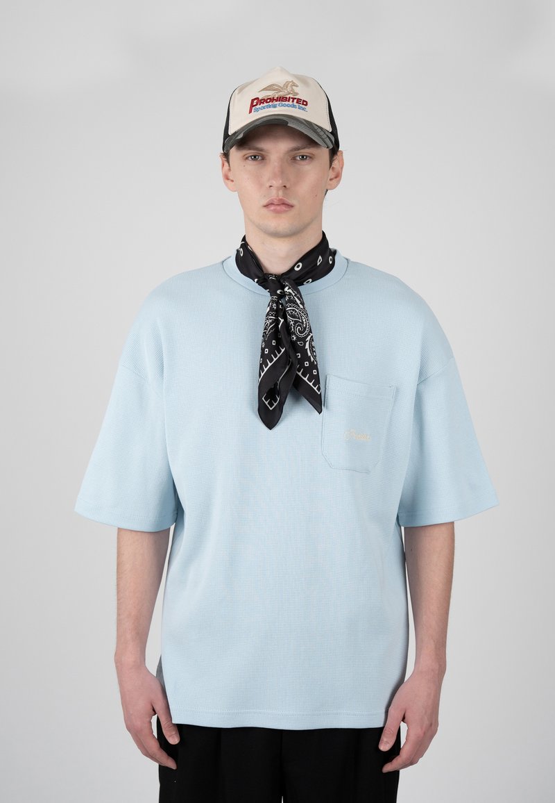 Jeune homme portant un t-shirt oversize bleu clair, une écharpe noire à motif paisley autour du cou, un pantalon noir et une casquette beige avec un texte rouge et bleu.