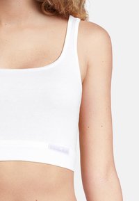Weißes Baumwoll-Crop-Top mit quadratischem Ausschnitt, dünnen Trägern und einem Markenetikett am Saum. Glatte Textur, taillierter Schnitt.