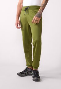 Pantalones jogger verde oliva con cintura elástica y cordón negro, con una textura suave y un diseño ajustado. Llevados con zapatillas gris-negras.