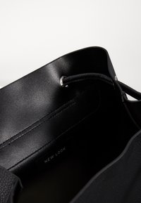 Wnętrze czarnej torby typu bucket z ekoskóry z okrągłym otworem, zaciąganym zamknięciem oraz widoczną metką marki. Teksturowane i gładkie powierzchnie.