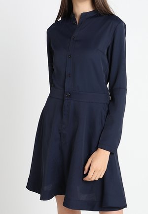 Nainen, joka käyttää laivastonsinistä, pitkähihnaista napitettavaa mekkoa, jossa on kaulus ja levenevä helma.