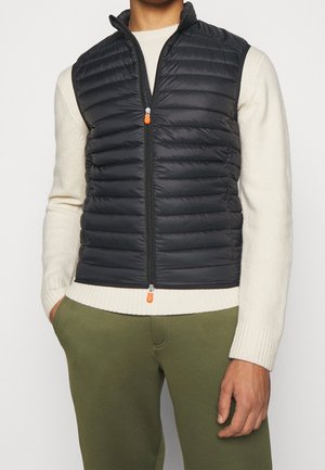 Homme portant un gilet matelassé noir sans manches sur un pull crème et un pantalon vert olive, debout avec une main dans la poche.